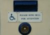 Close view of part of a closed door, showing a mail slot above two small signs, and below them, a buzzer. One sign is the universal icon for accessibility: a wheelchair. The other says, "Please ring bell for attention."