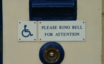 Close view of part of a closed door, showing a mail slot above two small signs, and below them, a buzzer. One sign is the universal icon for accessibility: a wheelchair. The other says, "Please ring bell for attention."