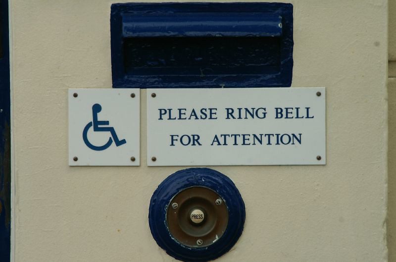 Close view of part of a closed door, showing a mail slot above two small signs, and below them, a buzzer. One sign is the universal icon for accessibility: a wheelchair. The other says, "Please ring bell for attention."