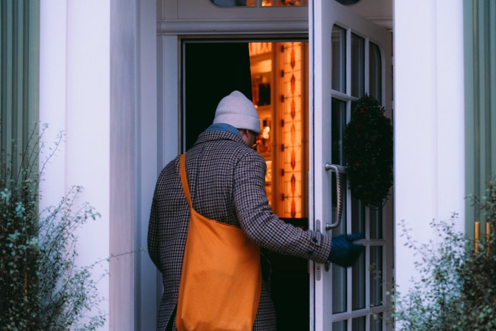 A three-quarters back view of a man in a brown/gray checked coat and a light gray watch cap entering an open door. To either side of the door are wispy shrubs, green against the white walls.His gloved right hand is on the door. He has an orange crossbody bag over his left shoulder. The room ahead of him, inside the house, has orange walls or the light gives an orangey glow.
