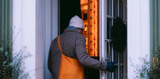 A back view of a man in a brown/gray checked coat and a light gray watch cap entering an open door. To either side of the door are wispy shrubs, green against the white walls.His gloved right hand is on the door. He has an orange crossbody bag over his left shoulder. The room ahead of him, inside the house, has orange walls or the light gives an orangey glow.