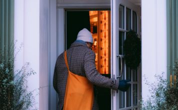 A back view of a man in a brown/gray checked coat and a light gray watch cap entering an open door. To either side of the door are wispy shrubs, green against the white walls.His gloved right hand is on the door. He has an orange crossbody bag over his left shoulder. The room ahead of him, inside the house, has orange walls or the light gives an orangey glow.