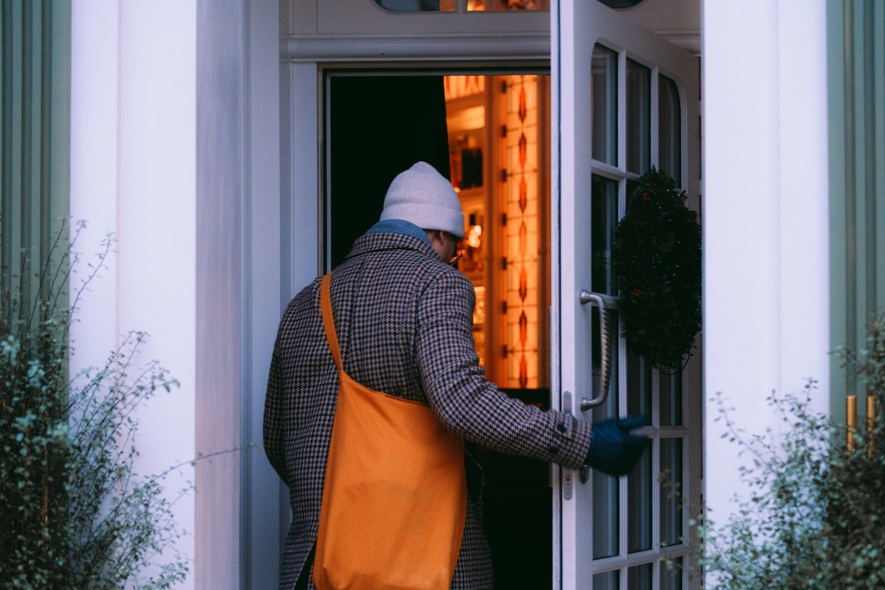 A back view of a man in a brown/gray checked coat and a light gray watch cap entering an open door. To either side of the door are wispy shrubs, green against the white walls.His gloved right hand is on the door. He has an orange crossbody bag over his left shoulder. The room ahead of him, inside the house, has orange walls or the light gives an orangey glow.