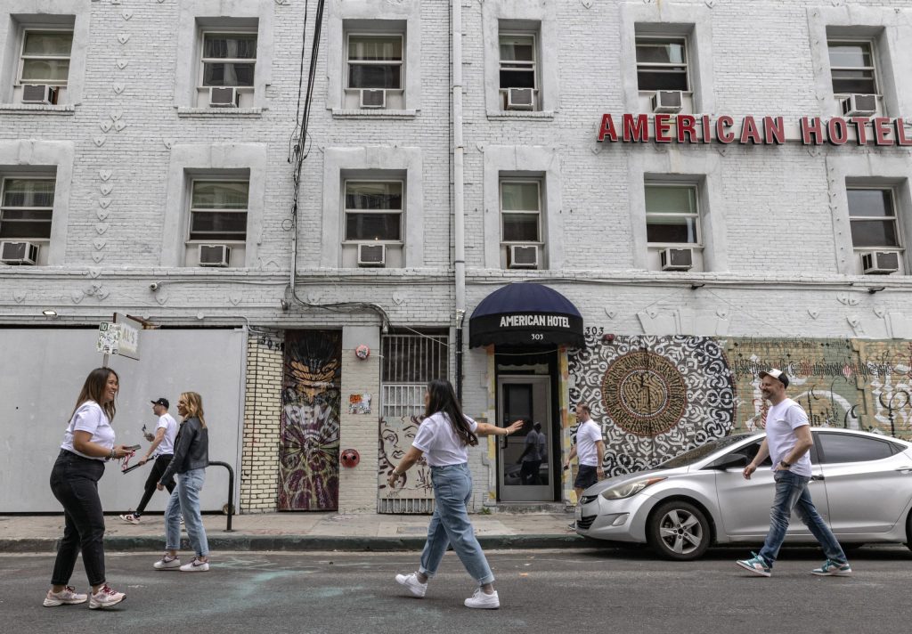 A gray brick building has a red sign that reads "American Hotel" in the front. Two women, both wearing white t-shirts and jeans, appear to be taking photos, but are in motion. On their right, a man with a cap, white t-shirt, and jeans walks by. Three other people are passing by in the background of the photo.