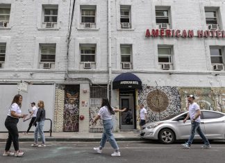 A gray brick building has a red sign that reads "American Hotel" in the front. Two women, both wearing white t-shirts and jeans, appear to be taking photos, but are in motion. On their right, a man with a cap, white t-shirt, and jeans walks by. Three other people are passing by in the background of the photo.