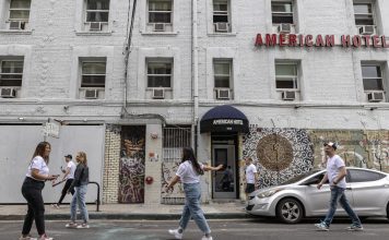 A gray brick building has a red sign that reads "American Hotel" in the front. Two women, both wearing white t-shirts and jeans, appear to be taking photos, but are in motion. On their right, a man with a cap, white t-shirt, and jeans walks by. Three other people are passing by in the background of the photo.