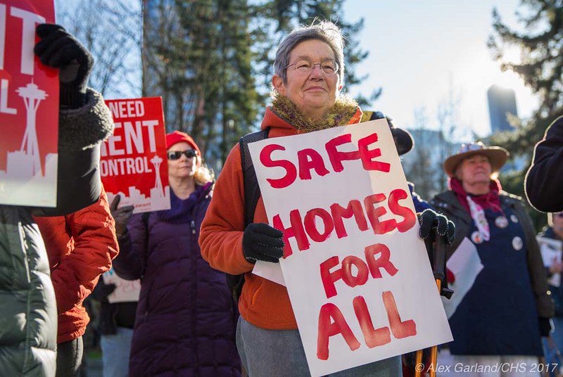 Several people in winter clothing stand outdoors on a sunny day holding signs. At center, a light-skinned woman in late middle age holds a sign that says "Safe Homes for All," written in red paint. Other people, partly visible, hold printed signs calling for rent control. Behind the tenant-protections protesters are hemlock trees and beyond them, partly visible, are tall buildings.