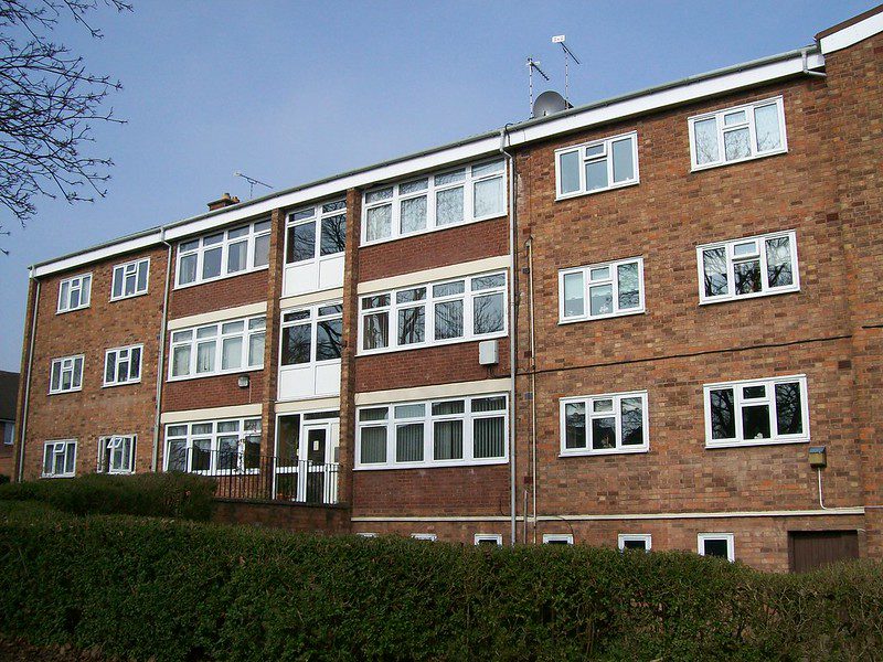 A three-story red/brown brick council housing building with white window frames. In the foreground, a thick green hedge. Behind the building, a clear blue sky. There are no people in the photo.