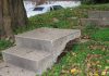 Concrete steps on a gently sloping grassy hill go up three steps, then become discontinuous with the steps above them. They're somehow (it's not clear how) raised up, so that on the third step a walker would have to step down before going on to the next step up.
