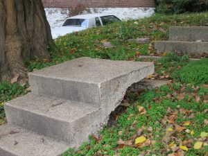 Concrete steps on a gently sloping grassy hill go up three steps, then become discontinuous with the steps above them. They're somehow (it's not clear how) raised up, so that on the third step a walker would have to step down before going on to the next step up.