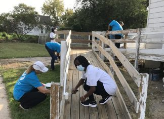 Five people are painting a wooden ramp leading up to a white house. Two are wearing white t-shirts and three are wearing blue t-shirts.