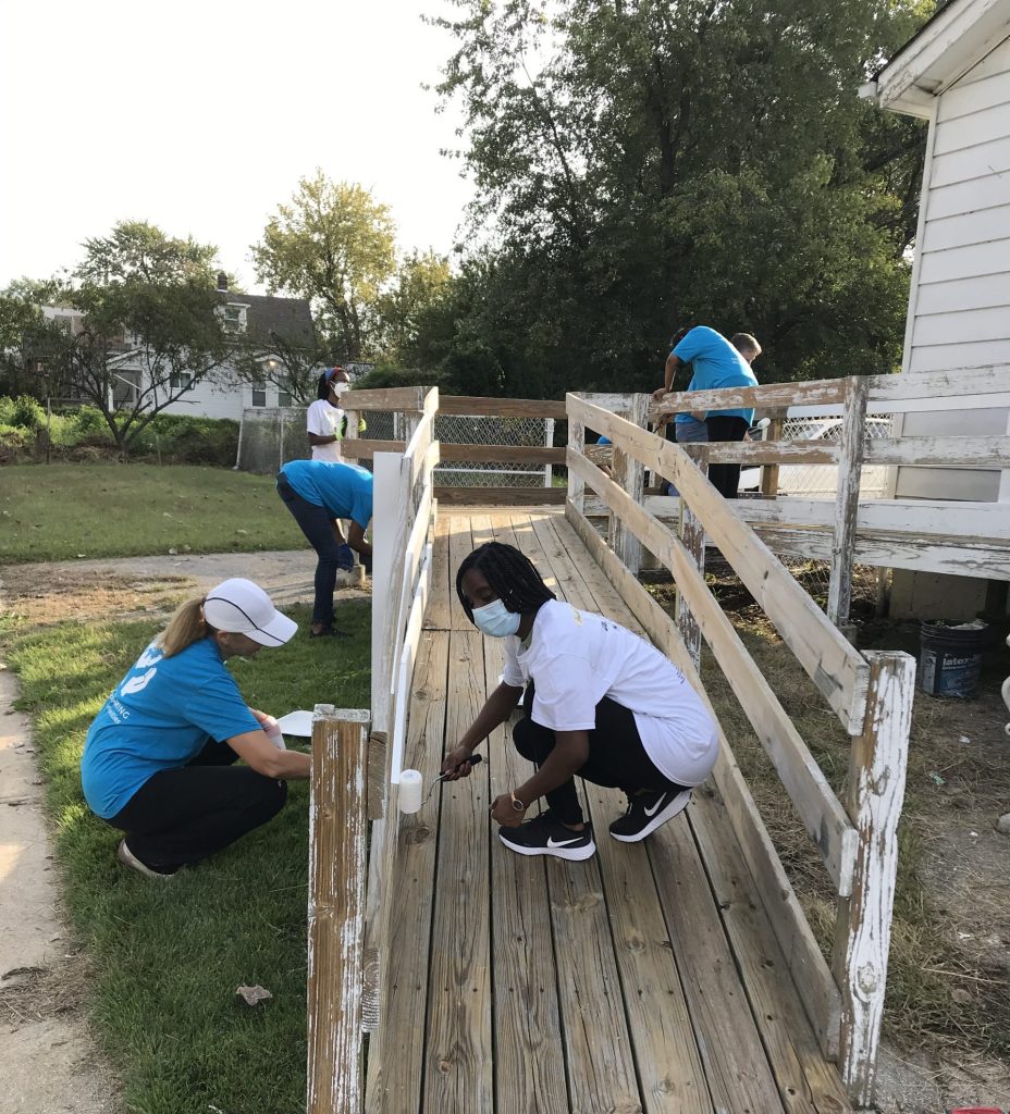 Five people are painting a wooden ramp leading up to a white house. Two are wearing white t-shirts and three are wearing blue t-shirts.