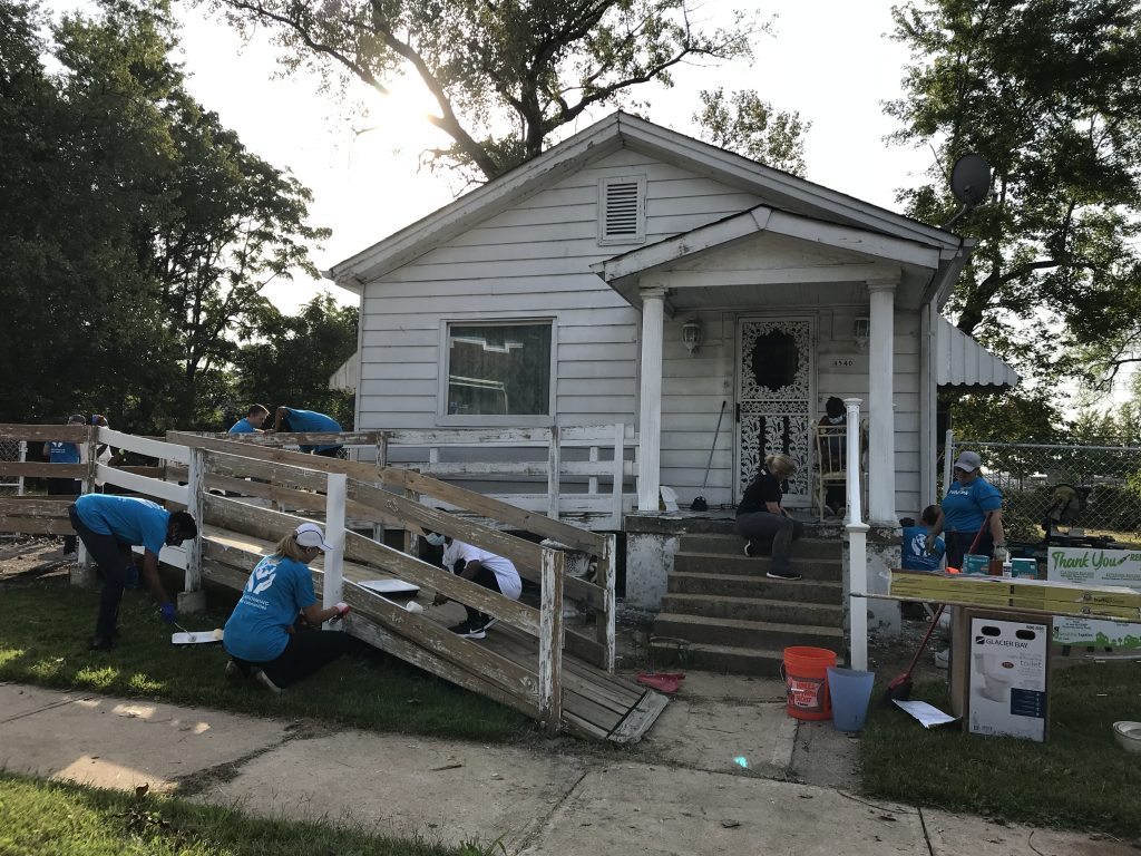 Workers, most wearing blue t-shirts, work on a white house. There is a wooden ramp in front that they are painting. The house is white and is surrounded by trees.