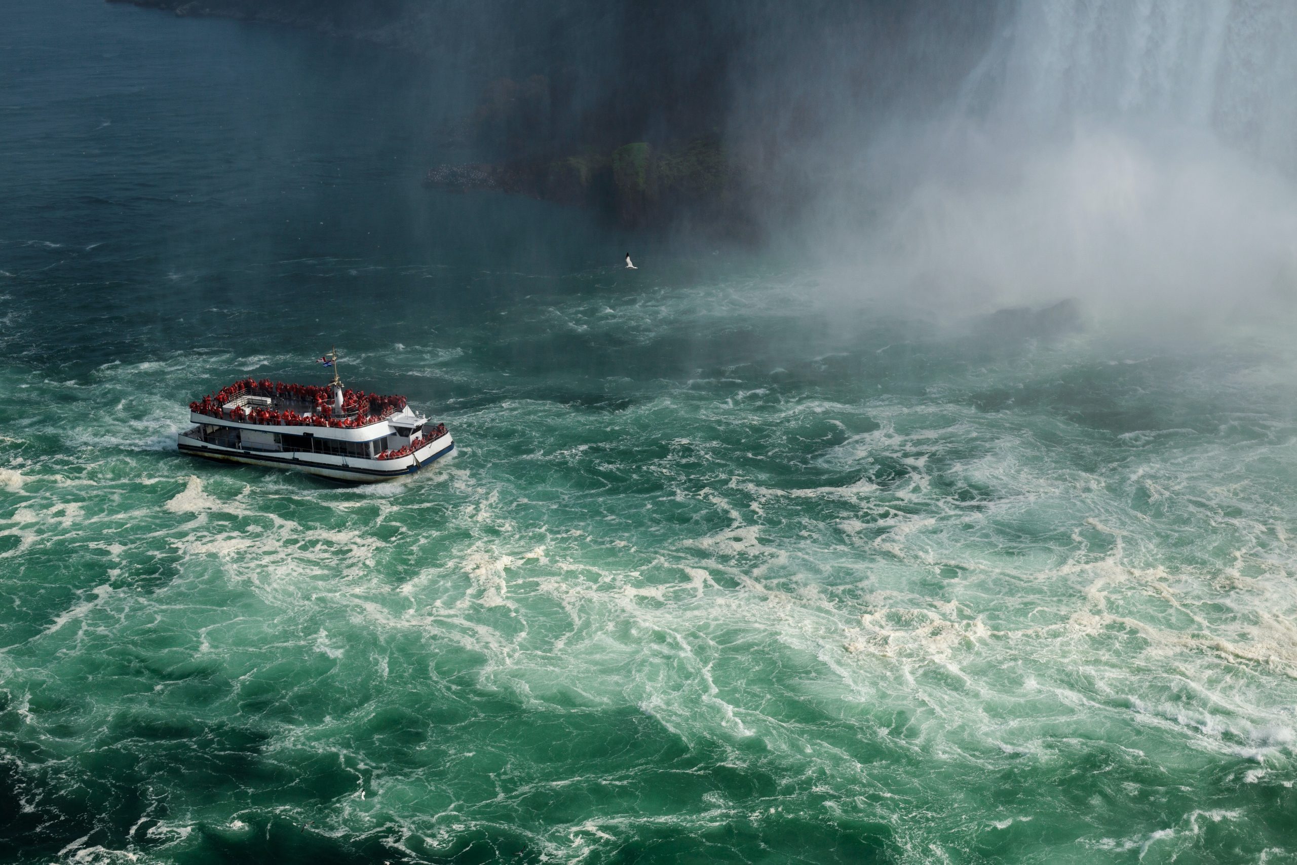View from above of a ferryboat on roiling waters, with clouds of mist off to the right. The water is blue-green with much white foam from the choppy sea