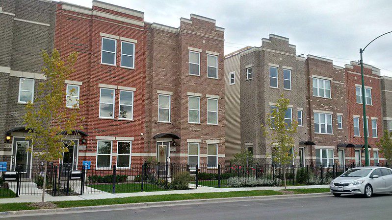 View from across the street of a row of six apartment buildings, all three stories, in varying brick shades. All have square patches of lawn in front and wrought-iron fences with gates. At far right is parked a silver sedan. There are no people in the photo.