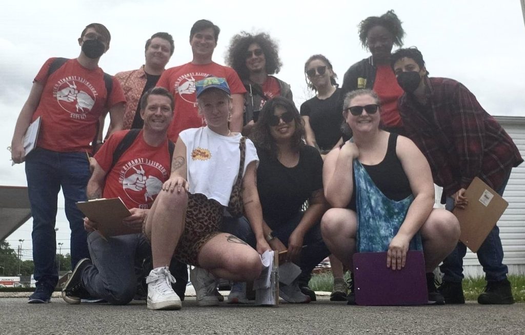 Eleven people, seven standing in back, four crouching in front, pose with clipboards. Several are wearing red T-shirts that say Louisville Tenants Union with an image of two hands fist-bumping. Several of the people are wearing masks; the ones who aren't are smiling.