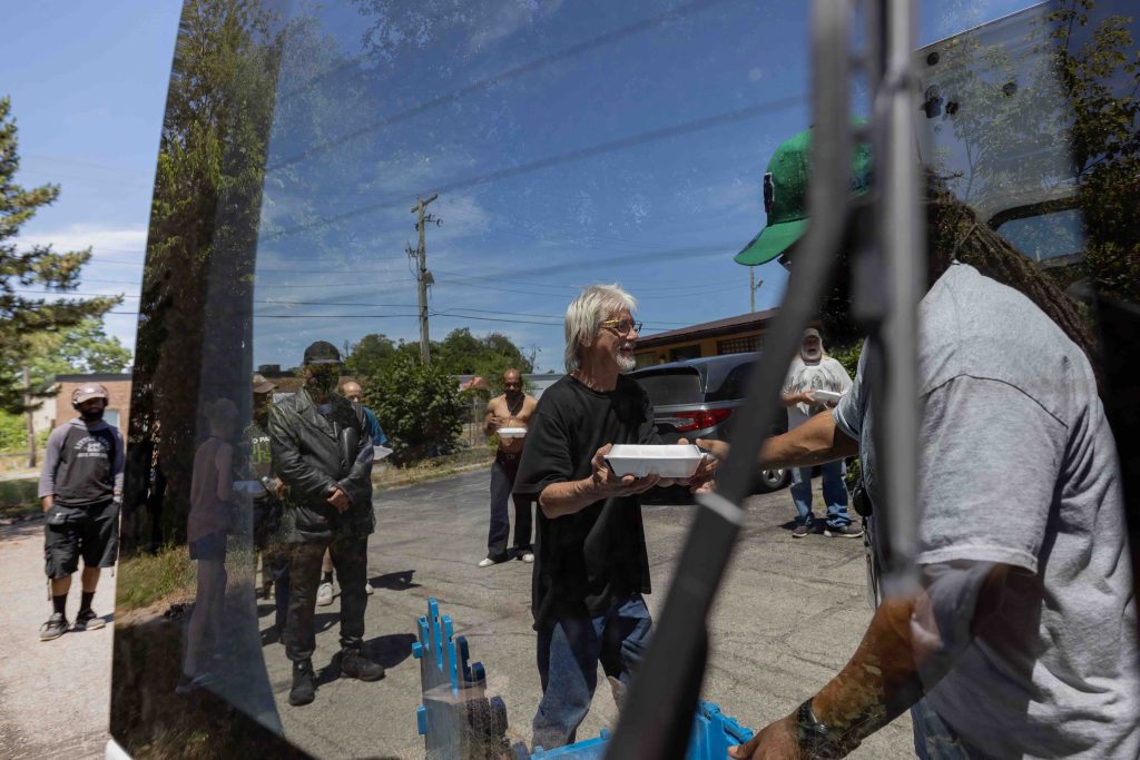 Seen mostly through a tinted car-door window, a man with dreads and wearing a green ball-cap hands a styrofoam meal container to a man with white hair. Several people are behind him in line.