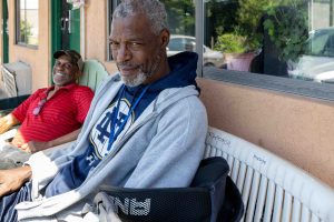 Two dark-skinned men with graying whiskers sit, one on a chair, one on a bench, in front of a peach-colored motel with green doors. At left, the man is wearing a bright red polo shirt and a ball cap, and is smiling broadly. The man on the bench is wearing a Notre Dame sweatshirt and looking relaxed but a bit dubious at having his photo taken.