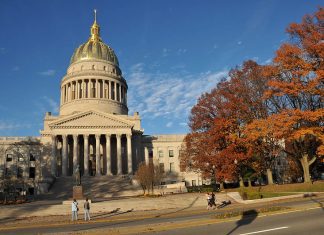An across-the-street view of a large domed building with broad steps leading up to the pillared front. Two people on the sidewalk are taking a photo, and two others are strolling by. The sky is a deep autumnal blue and the trees lining the plaza are in bright fall reds and oranges.