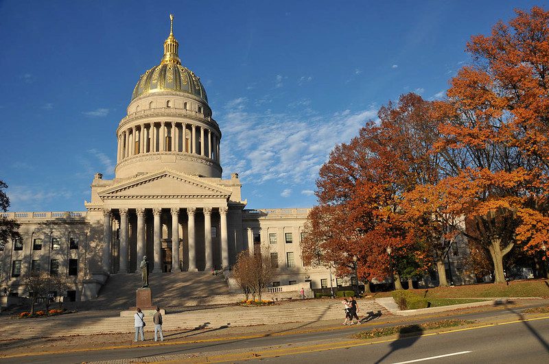 An across-the-street view of the West Virginia Capitol, a large domed building with broad steps leading up to the pillared front. Two people on the sidewalk are taking a photo, and two others are strolling by. The sky is a deep autumnal blue and the trees lining the plaza are in bright fall reds and oranges.