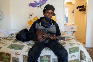 A young dark-skinned man sits on the edge of a bed holding a mandolin. He's wearing a ball cap turned sideway and a cloth mask is pulled down to show a smile. Behind him on the white wall is a mural of colorful insects and flowers.