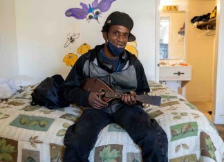 A young dark-skinned man sits on the edge of a bed holding a mandolin. He's wearing a ball cap turned sideway and a cloth mask is pulled down to show a smile. Behind him on the white wall is a mural of colorful insects and flowers.