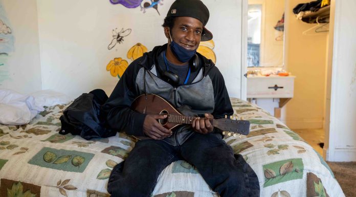A young dark-skinned man sits on the edge of a bed holding a mandolin. He's wearing a ball cap turned sideway and a cloth mask is pulled down to show a smile. Behind him on the white wall is a mural of colorful insects and flowers.