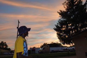 A dark-skinned bearded man in a brimmed hat and wearing a yellow T-shirt is silhouetted against a blue-and-pale orange sky late in the day. At right is a tall spruce tree, also in silhouette.