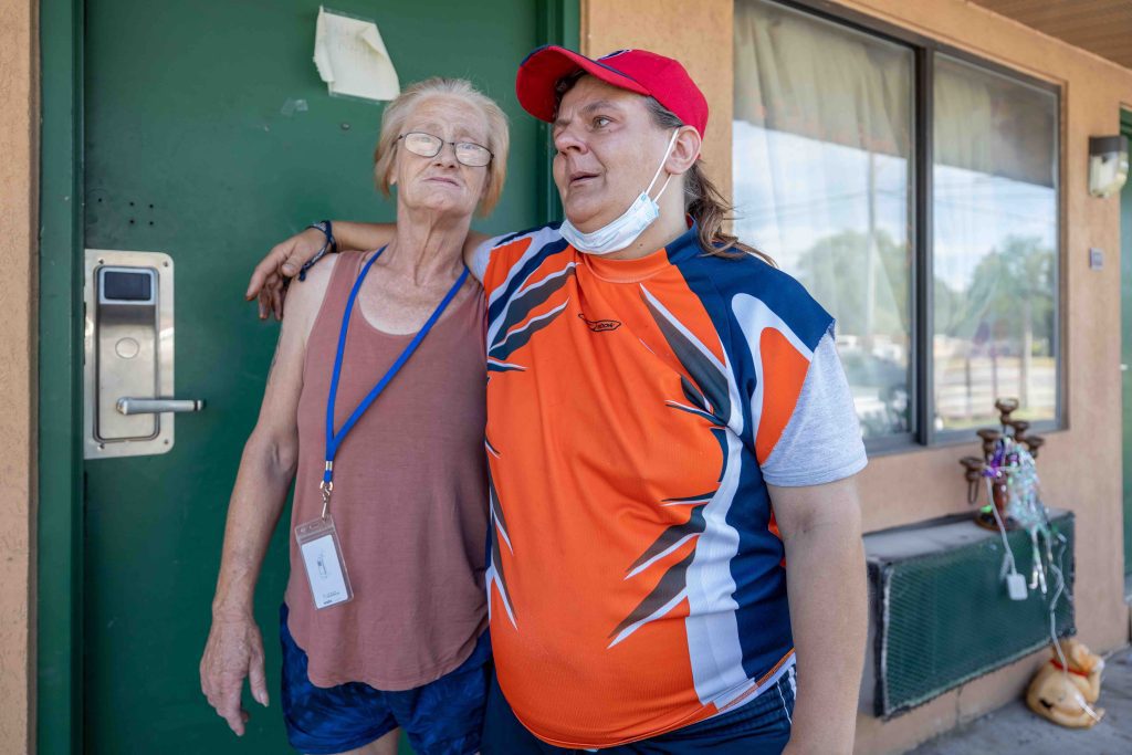 Two people stand by a green door in a peach-colored motel wall. At left, a woman with sandy hair and glasses has an ID tag on a lanyard around her neck. The person at right is in a bright orange and blue T shirt and has an arm around the woman.
