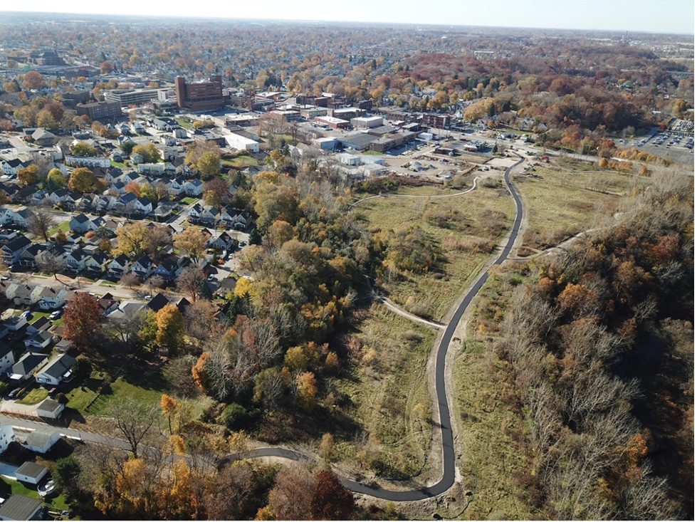 Drone view of a part of Cleveland's urban landscape in flat country, with a large grassy area running from top to bottom, with a narrow paved road traversing it. At left, the area is thickly built-up.