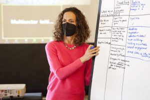 Woman wearing a mask and a pink shirt has one hand on a whiteboard. On the board, there is text written about the capital absorption framework.