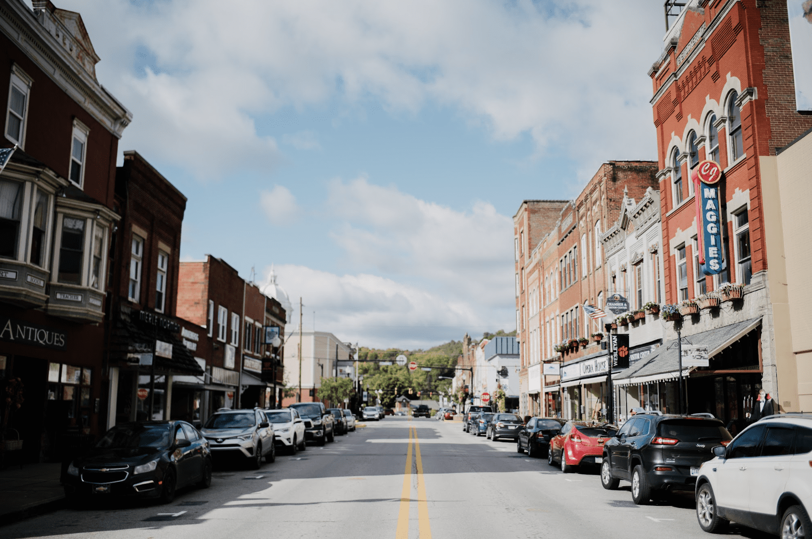 A town street seen from the center line, with cars parked along both sides, but no pedestrians visible. The street is lined with stores including an antique store, a building labeled "Maggies," and greenery in the distance. 