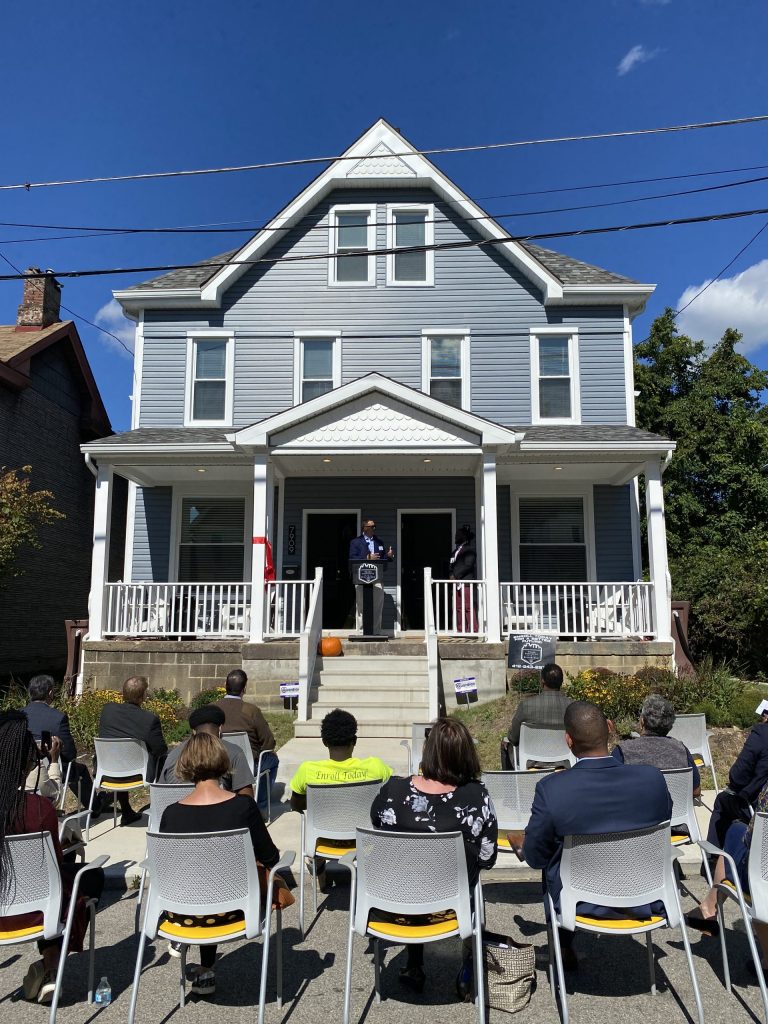 A view from the sidewalk of a 3-story gray house with wide front porch and steps down to the street. A wide red ribbon is tied to one of the columns supporting the porch roof. A man stands at a lectern on the porch, speaking to about a dozen people seated in stackable chairs, taking up part of the street. The audience is seen from the back.