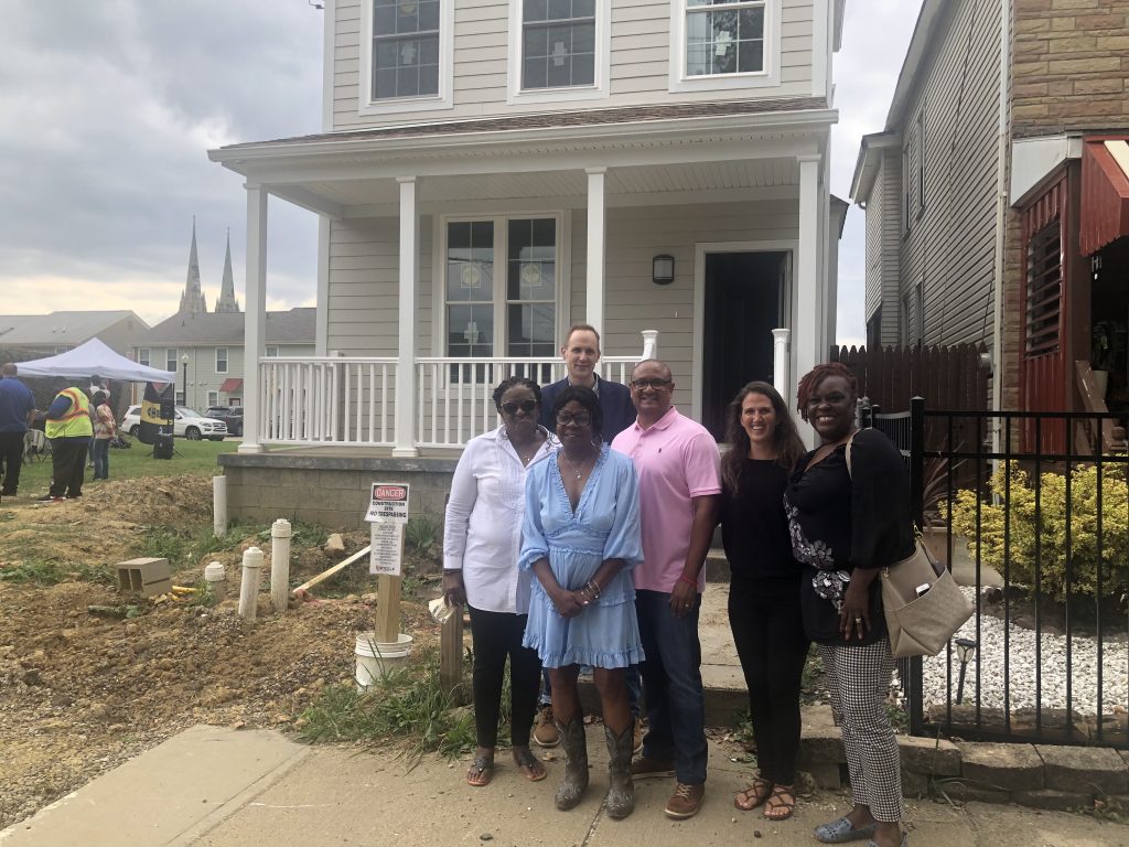 Two men and four women stand in front of a new-looking house, smiling for the camera. The front yard of the house looks dug up and there are pipes and a cinder block on the ground, adding to the new look. Beyond the house in the distance are other residential buildings and beyond them, twin spires of a church or cathedral. 