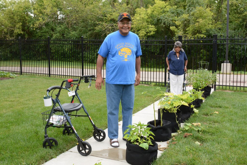 An outdoor view of an elderly Black man and woman standing on a walkway in a lawn. Lining the walk are large pots with vegetable plants. The woman, in a blue blouse and white slacks, and standing farther from the camera, has a walker right behind her. The man, closer to the camera, is standing by, but not holding onto, a walker. He's wearing a bright blue T-shirt with a yellow tree design, and is smiling broadly.