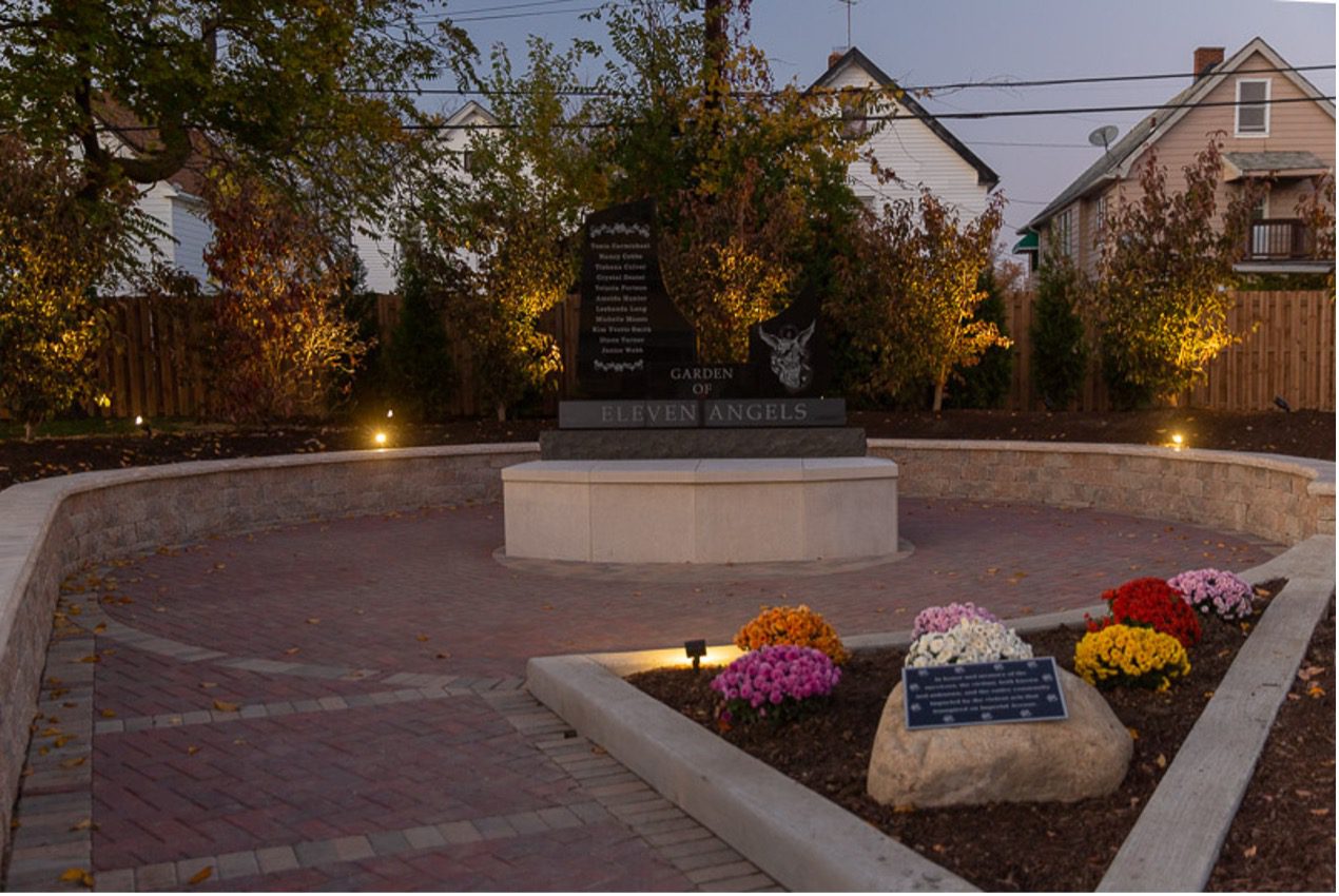 At dusk, a stone and brick memorial space, circular in shape with a central stone monument. Readable on the monument is the title "Eleven Angels." Small lights at the perimeter cast a gentle light on the space. The walkway leading to it is bordered with flower plantings and a large stone in the garden area bears another plaque (inscription too small to read). Behind the space are trees and shrubs and beyond them, a row of houses, only partly visible.