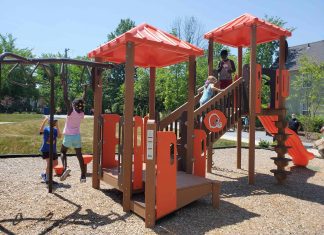 An orange and brown playground apparatus including a slide, monkey bars, and a treehouse, sits on a bed of wood chips in a grassy park on a sunny day. Four children of varying skin tones play on the equipment. Beyond the park area a man in uniform watches the playground and behind him is a clapboard house.