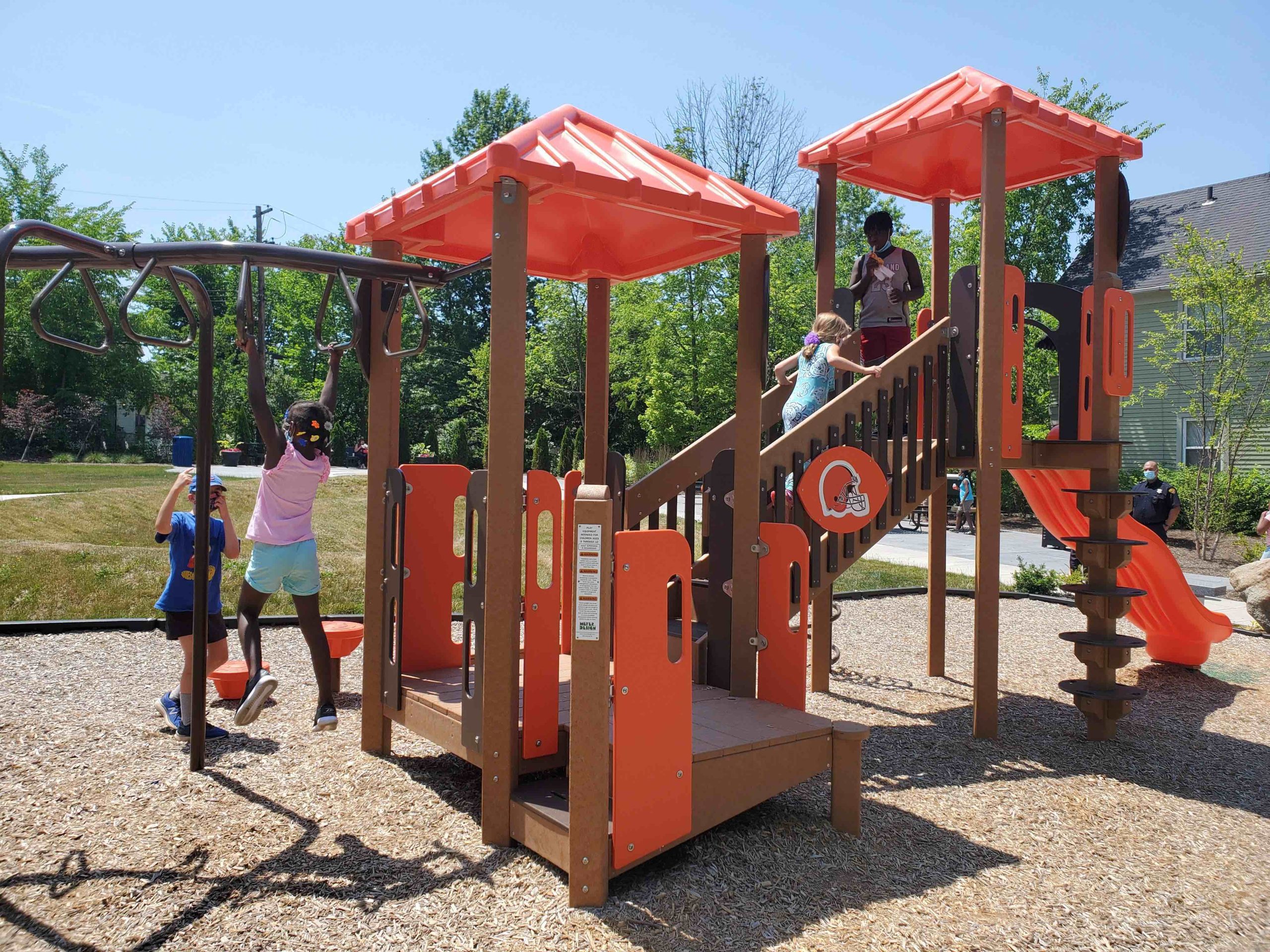 An orange and brown playground apparatus including a slide, monkey bars, and a treehouse, sits on a bed of wood chips in a grassy park on a sunny day. Four children of varying skin tones play on the equipment. Beyond the park area a man in uniform watches the playground and behind him is a clapboard house.