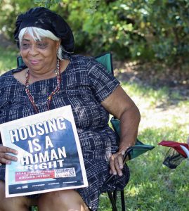 A middle-aged white-haired Black woman, wearing a black and white tweed-pattern dress and wearing a floppy beret, is sitting in a collapsible chair in a grassy area with shrubs behind her. She holds a sign reading "Housing is a Human Right." She is looking off to her right with a half-smile. St. Petersburg, Florida