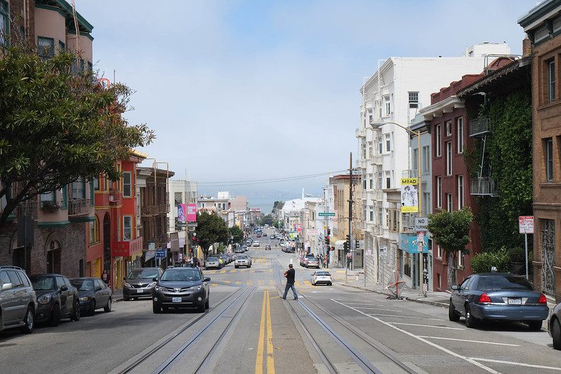 A downhill view from the center yellow line of a street in San Francisco, taking in several blocks. Several cars are parked along the left, uphill side. Fewer cars are parked on the right side. A man in dark clothes is crossing from right. The sky behind the scene is light blue and cloudy white.