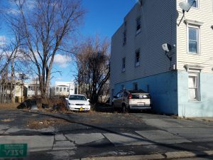 A derelict lot on a city street in winter or early spring; the trees beyond the lot are bare. Two cars are parked there, and rubbish or broken parts of things lie in the tall grass. Part of the lot is paved with cracked asphalt. The street beyond this one is visible, with two-story residential buildings. The sky is bright blue with small clouds in the distance.