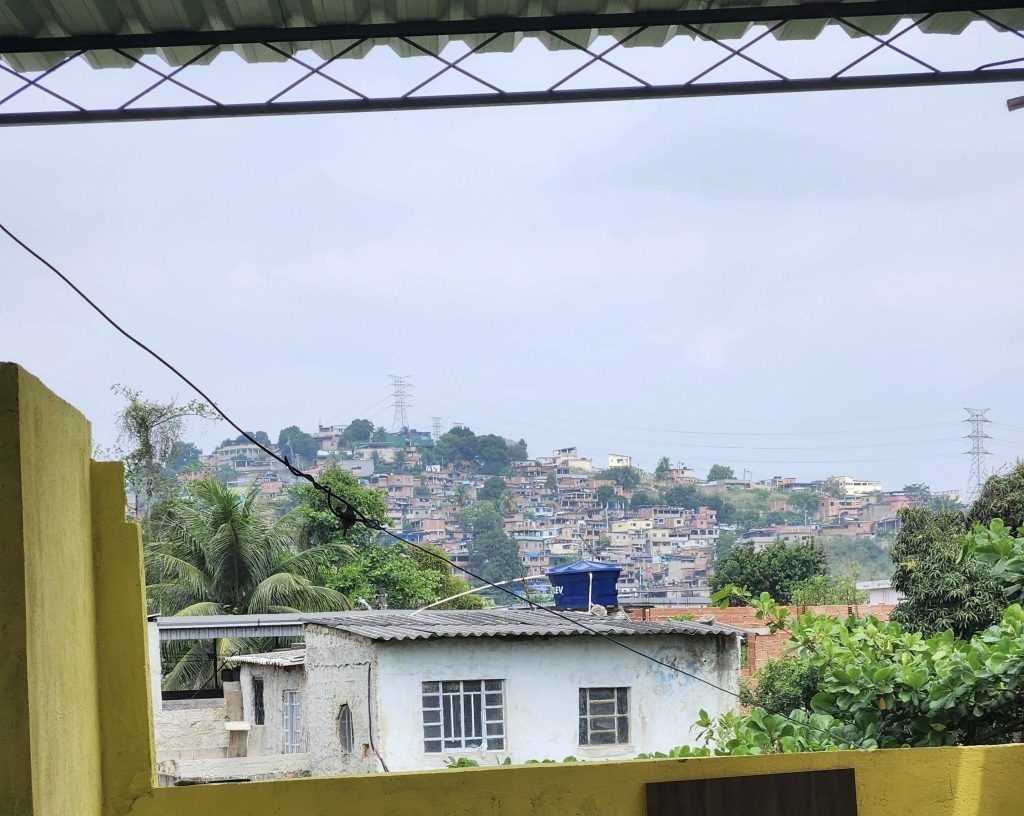 A favela of Rio de Janeiro. In the foreground is a small white building with a corrugated tin roof. Beyond it, in the distance, is a hilly landscape covered with similar dwellings. Tall power lines are visible in the distance.