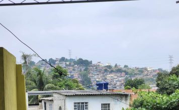 A favela of Rio de Janeiro. In the foreground is a small white building with a corrugated tin roof. Beyond it, in the distance, is a hilly landscape covered with similar dwellings. Tall power lines are visible in the distance.