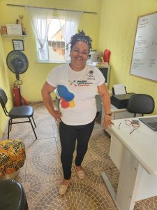 A light-brown skinned woman stands in a yellow room, smiling broadly. Her braided hair is pulled back, and she is wearing black leggings and a white T-shirt that has an abstract design in primary colors and is printed "Painel Unificador Favelas." One hand is on her hip, the other on a white desk.