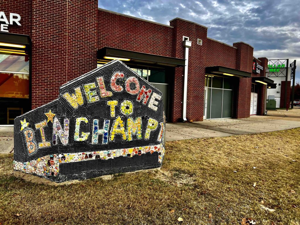 On a lawn in front of a brick building with large glass panels that look as if they might have been garage bays at one point, stands a welcome sign made of mosaic tile. The underlying structure is invisible but may be concrete and forms a boxy semi-circle with a peak. The mosaic tiles are small and spell out "Welcome to Binghampton" in varied colors on a background of tiny black tiles. There are two stars over the letters B and I, and near the bottom is a band of freeform mosaics depicting faces. They're very small and hard to discern individually.