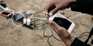 View of brown-skinned hands holding a cellphone with charging cable attached. On the ground nearby are power strips with several other phones being charge.