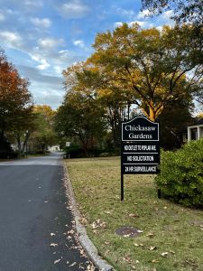 A road in a neighborhood (no cars or people to be seen) with grass, shrubs, and trees lining it. The sky is blue with some clouds. A sign posted by the road, in white lettering on a black background, says "Chicasaw Gardens/No outlet to Poplar Ave./No solicitation/24 hr surveillance."