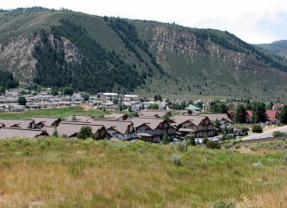 A view of a city from a grassy hillside. Just beyond the hill is a large settlement of one- and two-story houses, mostly visible by their roofs. Beyond them rises a steep, evergreen-dotted, and rocky mountain. The sky far beyond is white, except for a bit of blue at the right. No people are visible in this photo.