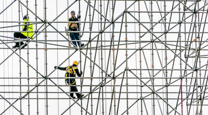 Three men in work clothes including helmets and tool belts stand on scaffolding. It looks as though the scaffolding is standing alone but it is erected against a white building, creating that effect.