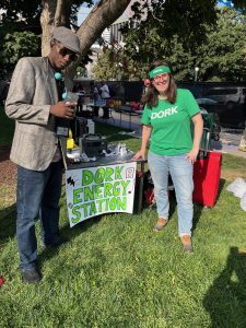 Two people stand on either side of a table set up in a public area. Taped to the table is a sign reading "Dork Energy Station" and on it are several cables and adapters to charge phones with. On the left is a young dark-skinned man in a gray tweed blazer and soft cap holding a cellphone and wearing sunglasses. At right is a white woman in jeans and green T-shirt (which says Dork). She has a green headband, wears glasses, and is smiling at the camera.